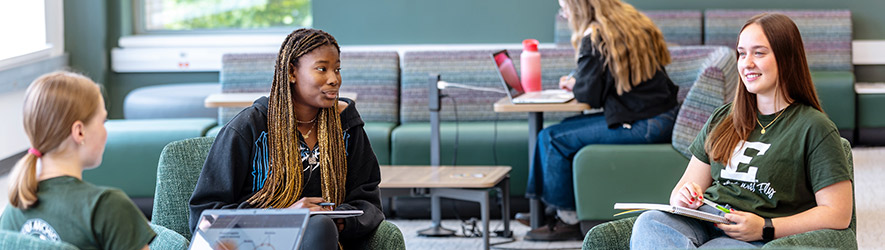 Three college students sit in comfortable armchairs talking and studying in a modern campus lounge, while another student works on a laptop in the background.